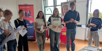 Schüler*innen stehen in einem Klassenzimmer und halten Informationsmaterial in den Händen